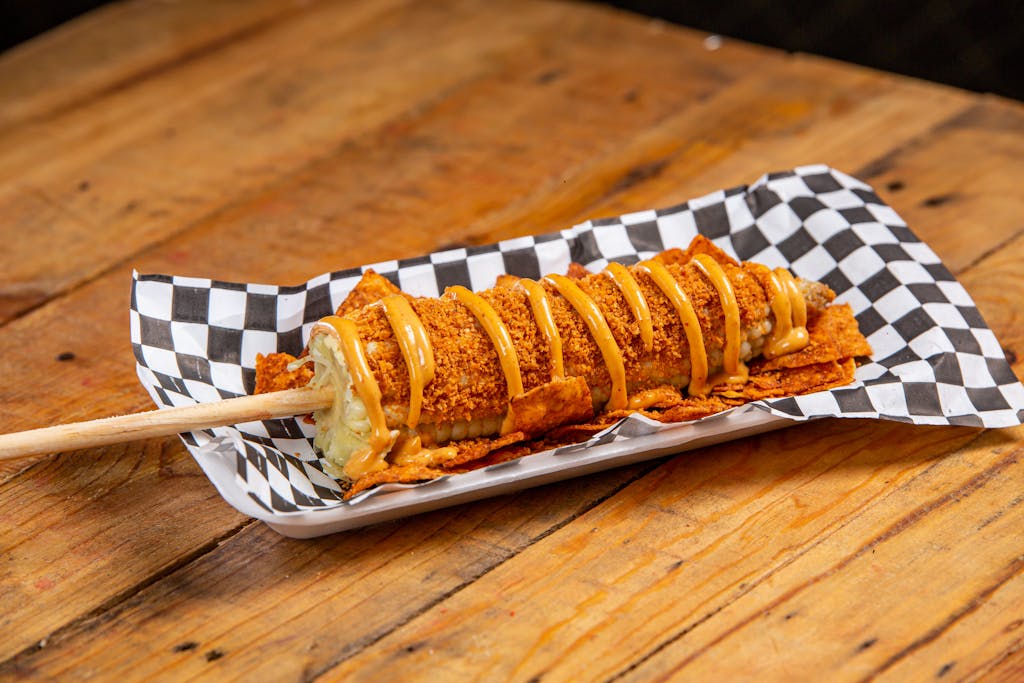 A delicious crispy corn dog drizzled with mustard on a checkered paper stand.